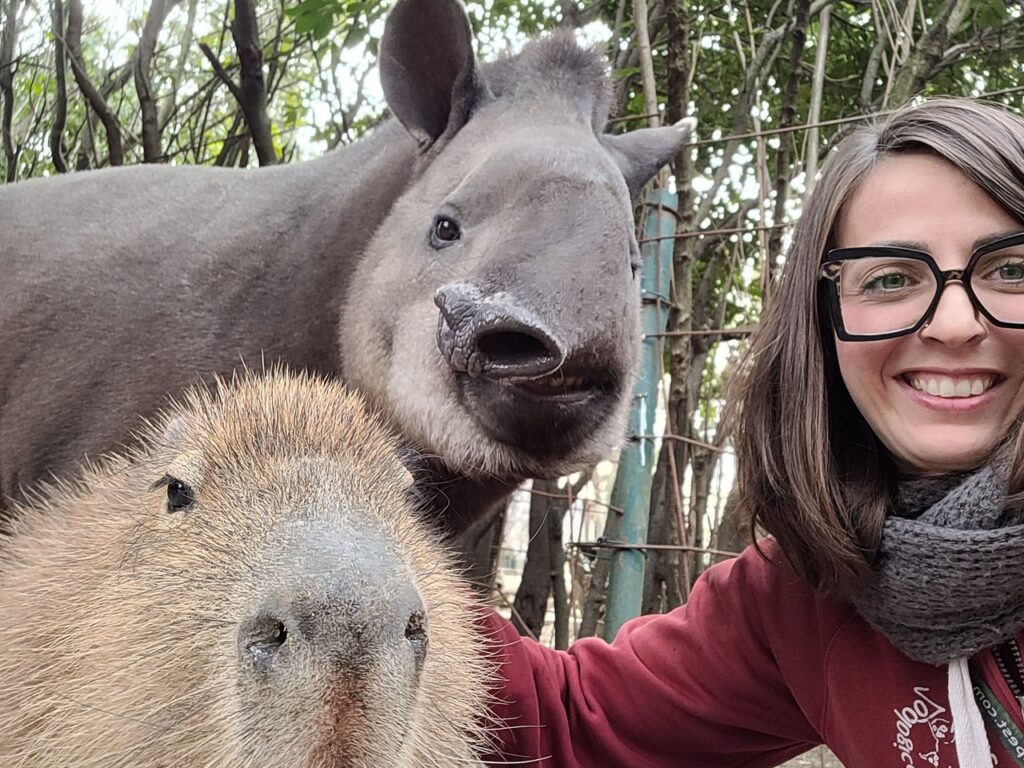 La dott.ssa Rossana Cordon dell'Ospedale Veterinario Ardenza sorride mentre scatta un selfie insieme a un capibara e un tapiro, in un ambiente esterno con vegetazione e recinzioni sullo sfondo.